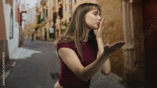 Fotografía Young woman blows a kiss with hand raised while standing in a narrow old town street, cropped midriff visible and casual pose; playful affection
