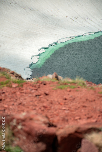 Glacial Mountain Lake Shoreline Showing Melting Glacier Ice, Red Volcanic Soil, and Deep Blue Water