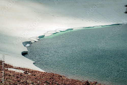 Glacial Mountain Lake Shoreline Showing Melting Glacier Ice, Red Volcanic Soil, and Deep Blue Water