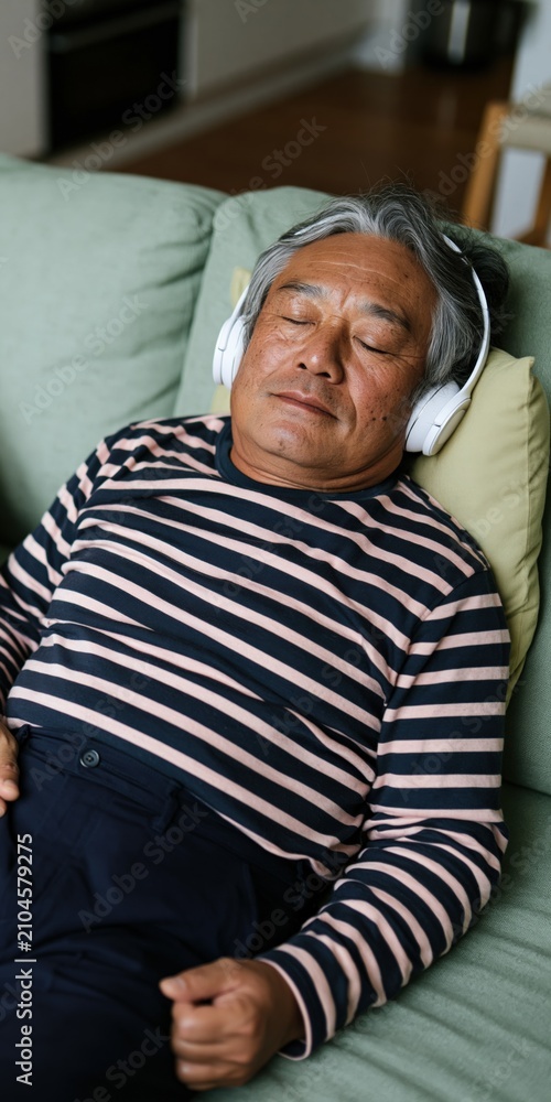 Fototapeta premium Older man resting on couch with headphones and striped shirt eyes closed for relaxation background and sleep therapy design