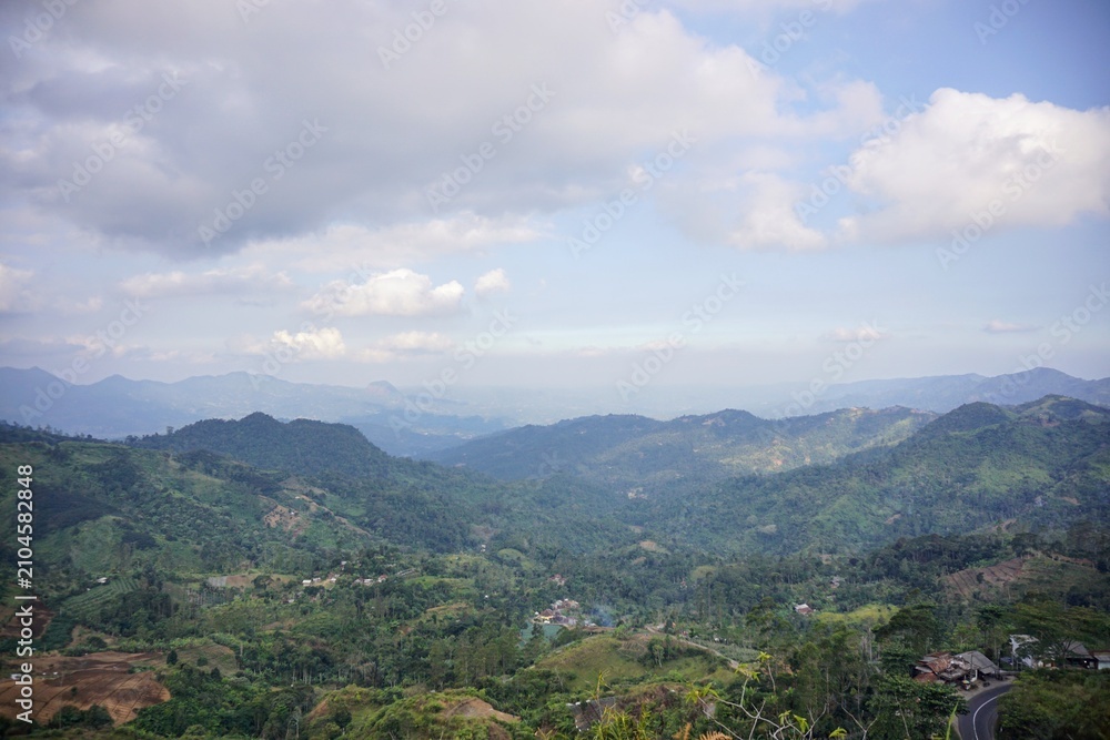 Obraz premium Panoramic scenic view of vast green hills with a majestic mountain range background under a bright cloudy blue sky.