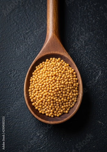 A high-angle close-up shot of golden yellow mustard seeds piled inside a rustic wooden spoon, resting on a dark, textured stone tabletop, texture, overhead, spoon