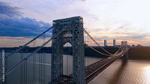 Heading to the top of the massive metal support of the George Washington Bridge at sunset. Lots of traffic on the structure. Boat is moving by the river.