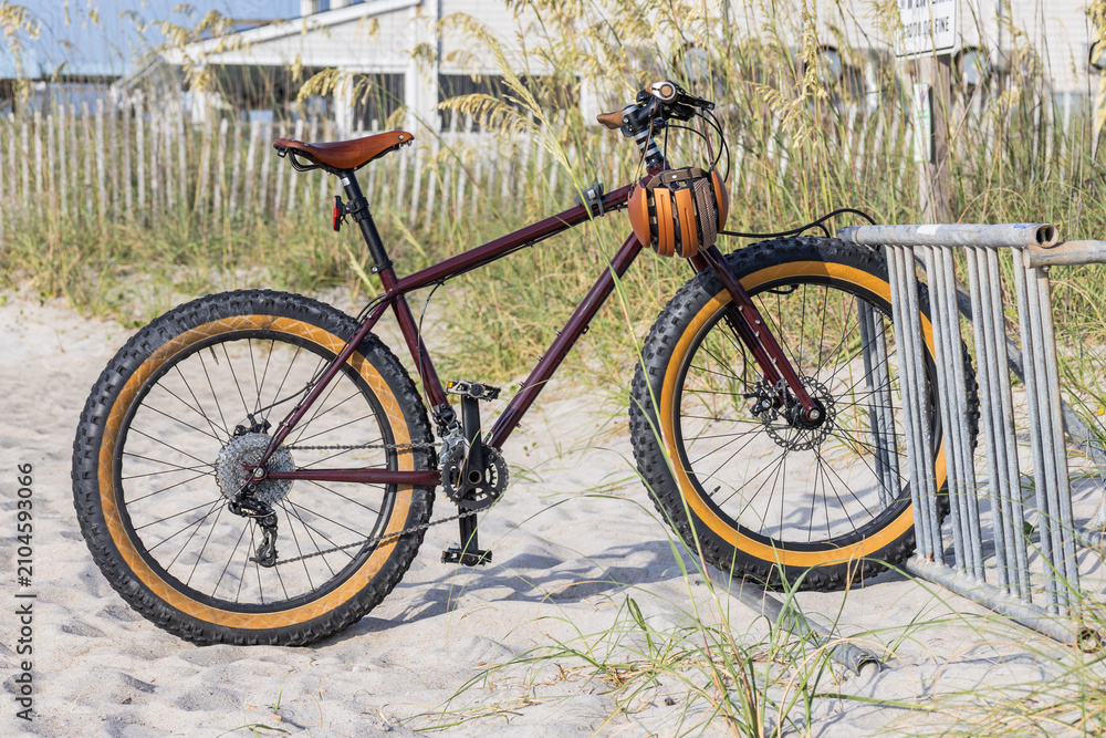 Fototapeta premium Sturdy bicycle rests on sandy beach beside wooden fence. Thick tires suited for rough terrain. Rustic brown frame and seat contrast with golden wheels. Natural light highlights texture of sand