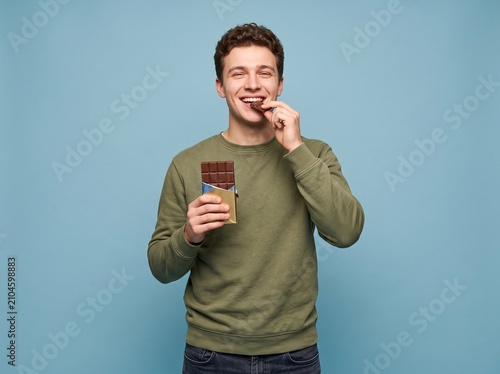 Happy Young Man Enjoying Delicious Chocolate Bar on Blue Background