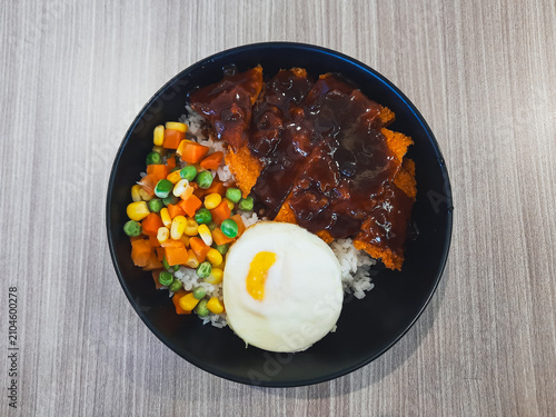 Rice bowl chicken katsu. Black bowl holds steamed rice, colorful veggies, and fried chicken chop. Sunny side up egg rests beside the savory centerpiece. Wooden table, tempting, restaurant menu