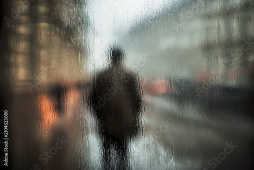 Silhouetted figure walking down a rainy city street seen through a wet glass window with blurred urban buildings and glowing lights at dusk