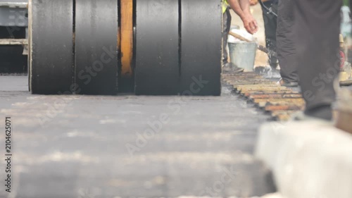 Black road roller presses the pavement surface. Worker in dark pants walks beside the machine. Concrete blocks mark the road's side. Truck stands in the distance