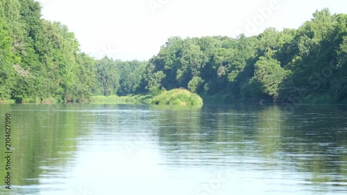 Serene river reflects sunlight. Calm water flows through forest. Dense green trees line both banks. Lush grassy patches grow in the river bend. Peaceful natural setting for calm moments