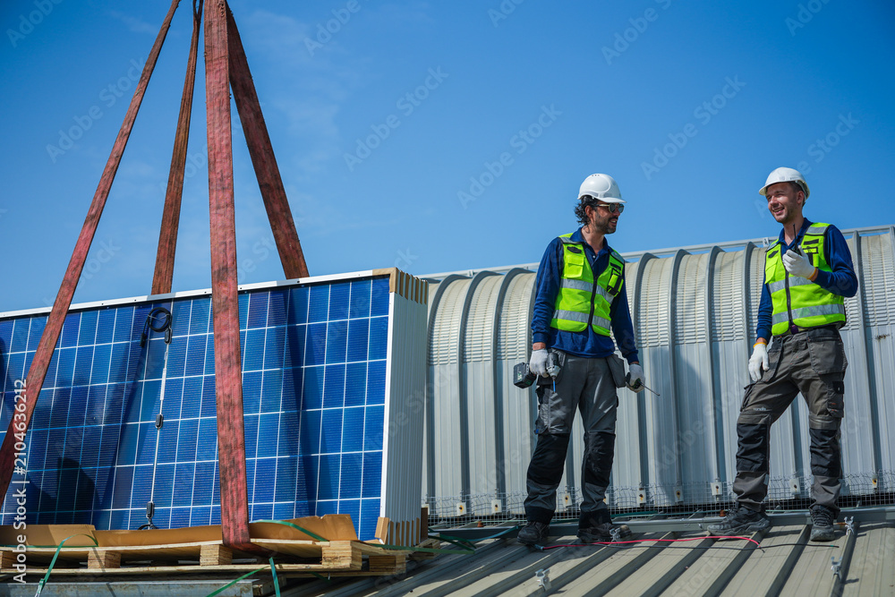 Obraz premium Two renewable energy specialists in hard hats and hi vis workwear steadily discuss panel distribution beside the delivery stack high on an industrial warehouse roof in a Thai city.