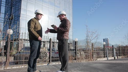 Two construction professionals handshake and consult with tablet in front of reflective glass office tower under clear blue sky