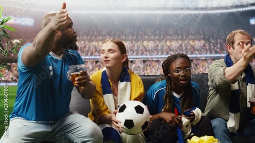 Tight framing shows football supporters with beer cups and ball in hands watching match broadcast as if inside the stadium while soccer fans drink alcohol and eat chips at home during stadium viewing.