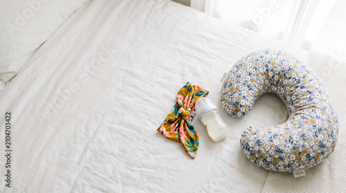 Cozy setup for first-time parents advertising motherhood essentials featuring a soft nursing pillow baby bottle and floral hair accessory on a white bed