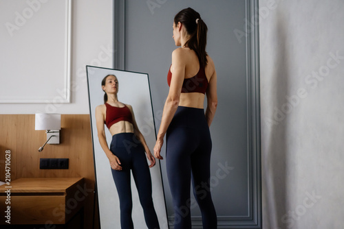 Woman standing in sportswear, looking at her reflection in a full length mirror, focusing on her appearance and portraying issues with body image and eating disorders