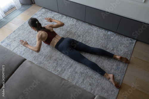 Woman lying on a rug in a modern living room, performing a back muscles exercise, focusing on strength training and maintaining a fit and healthy lifestyle with a home workout