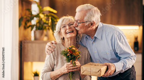A loving senior man kisses his smiling wife on the cheek while she holds a bouquet of flowers and a gift, celebrating a special occasion at home.