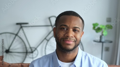 Close-up portrait of a young, confident, handsome african american man looking at the camera and smiling, sitting on a sofa in his modern living room. Happy millennial guy posing at home