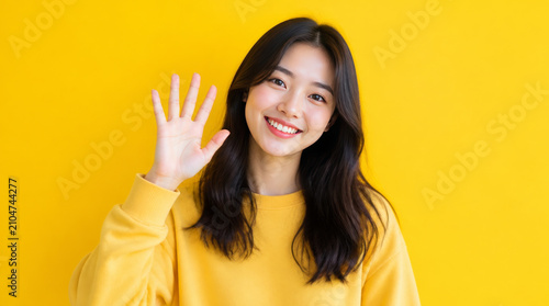Cheerful young woman waving hello against bright yellow background, expressing joy and friendliness