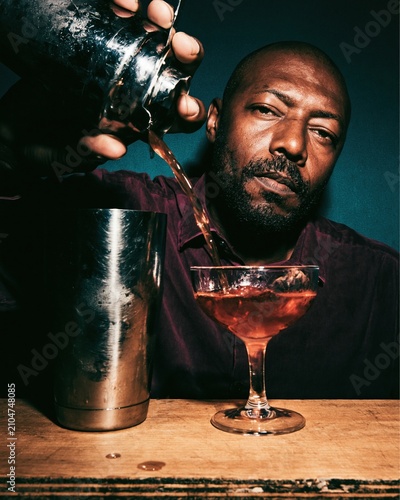 Serene man straining a chilled cocktail from a metal shaker into a glass for a moody drink menu, featuring dramatic rim lighting and copy space
