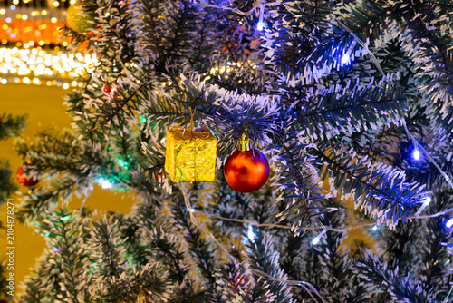 Red ball and gold gift box hanging on christmas tree branch close up with white blue led fairy lights illumination display at night