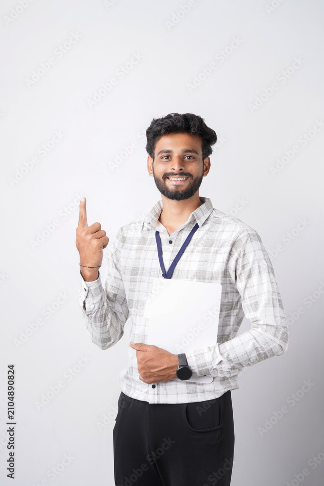 Fototapeta premium A smiling young man in a checkered shirt points upwards with his index finger against a plain white studio background.