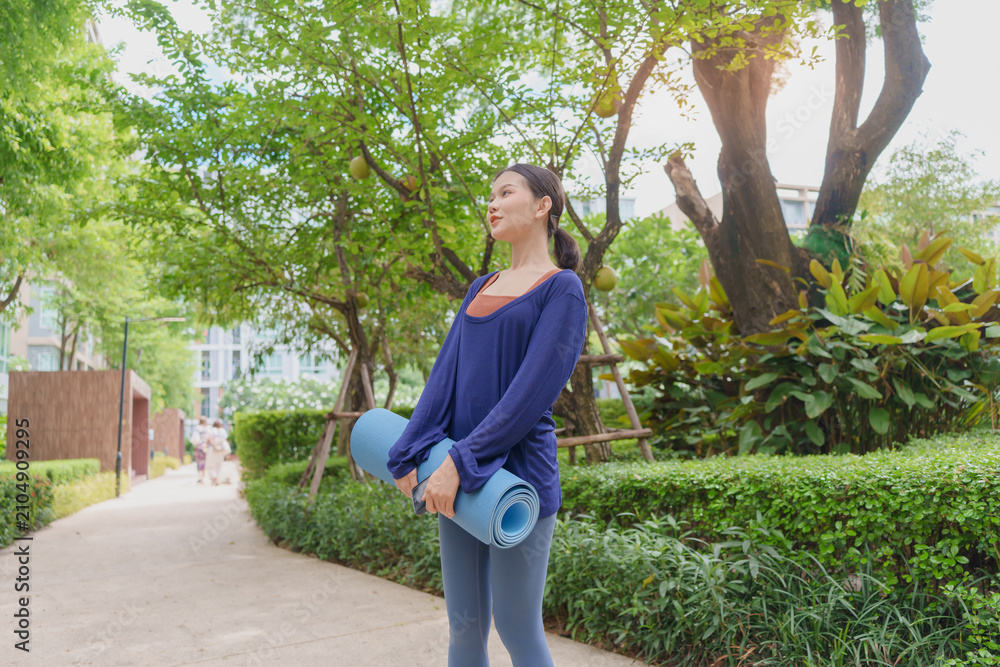 Fototapeta premium Asian Gen Z woman carrying yoga mat walking to park for wellness healthy lifestyle mindfulness and outdoor exercise.
