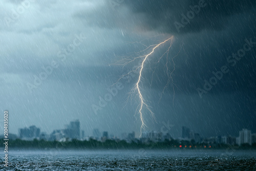 Thunder crackles above a stormy city skyline. Rain streaks the dark, swirling sky. Brilliant flash illuminates the distant buildings. Urban sprawl reflects in the turbulent waters