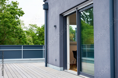 Sliding glass door stands open between composite deck terrace and modern apartment interior with gray stucco facade, metal railing, and green trees surround empty rooftop terrace under overcast sky