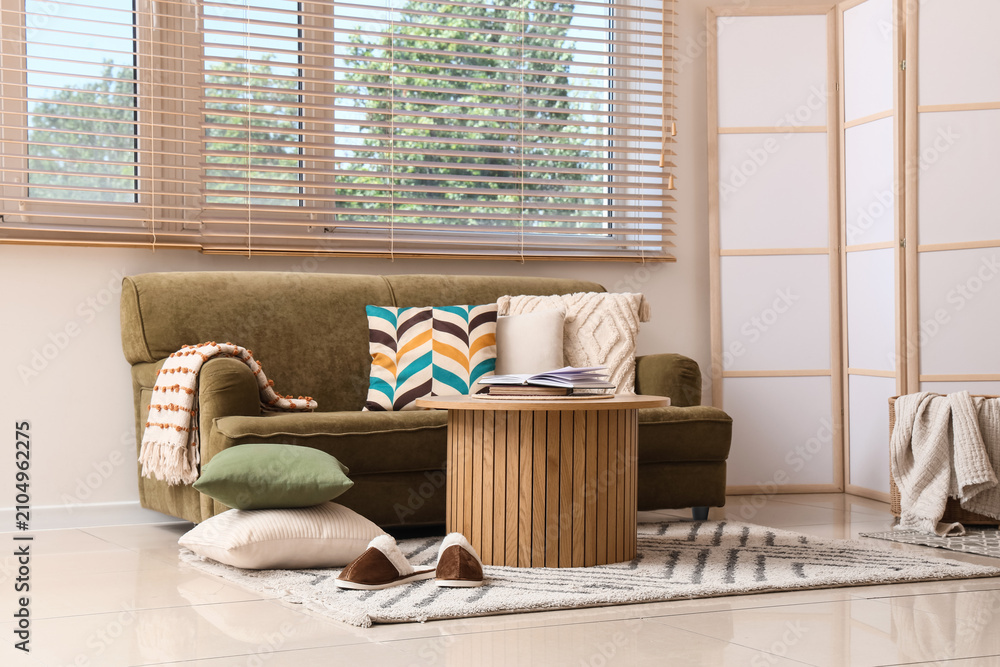 Obraz premium Cozy living room interior with a comfortable olive green sofa, wooden coffee table, and window blinds