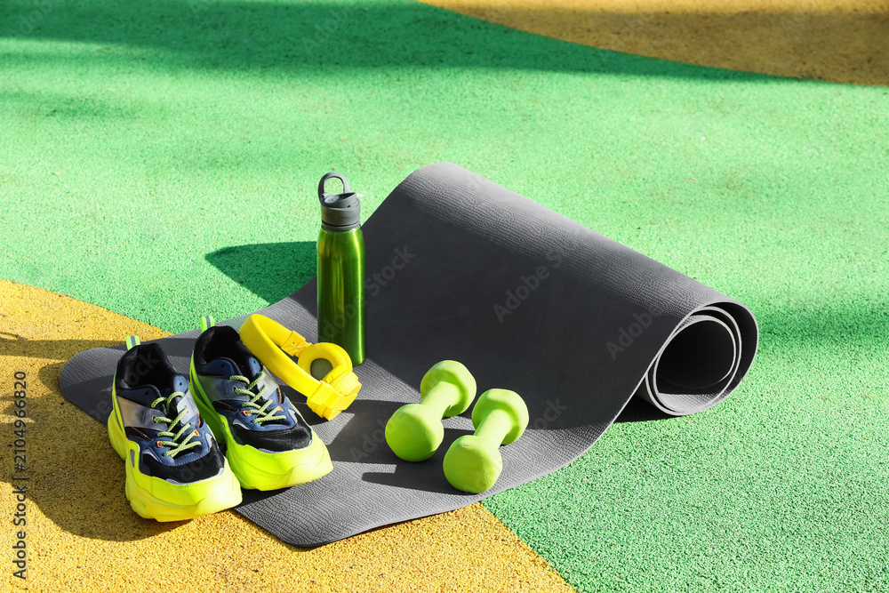 Fototapeta premium Fitness mat with sneakers, headphones, water bottle and dumbbells on sports ground