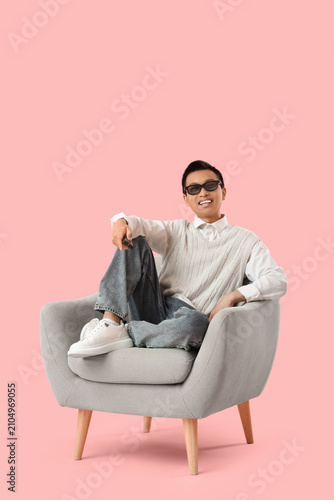 Young Asian man with TV remote controller sitting on armchair against pink ba...