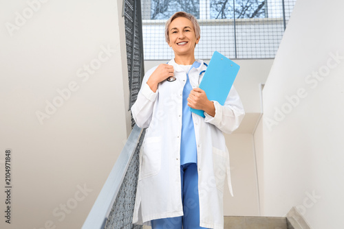 Happy mature female doctor with clipboard and stethoscope on stairs in clinic