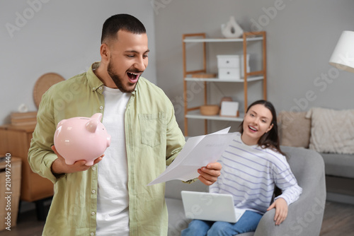 Happy young man with refund form and piggy bank at home
