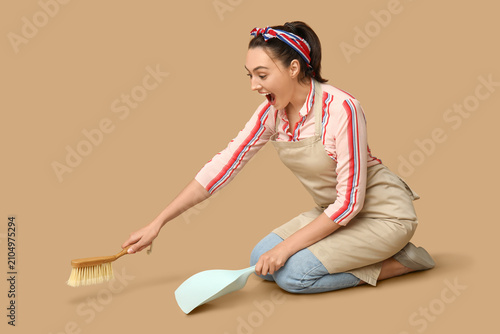 Portrait of happy young housewife in apron with dustpan and brush on brown ba...