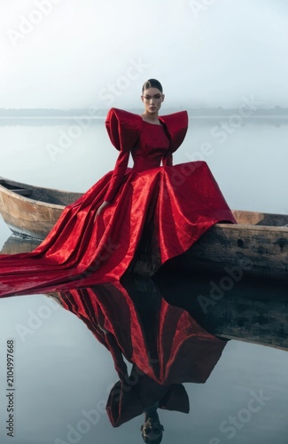 High fashion editorial portrait of a woman in an extravagant red voluminous gown sitting in a rustic wooden boat on a misty calm lake with dramatic mirror water reflection