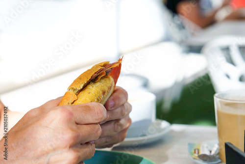 Hands holding a freshly made baguette sandwich with serrano ham, enjoying breakfast or a snack at an outdoor cafe