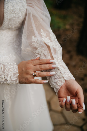 The bride in a white wedding dress embroidered with various patterns and beads holds her sleeve with her hand, showing off her manicure and gold wedding ring on her finger