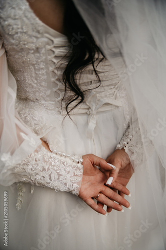 The bride, her back turned, in a white wedding dress embroidered with various patterns and beads with a long transparent veil, folded her palms together