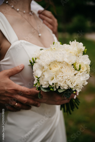 The bride in a white, tight, open dress holds a bouquet of white roses in front of her, a precious jewel around her neck, her groom hugs her from behind