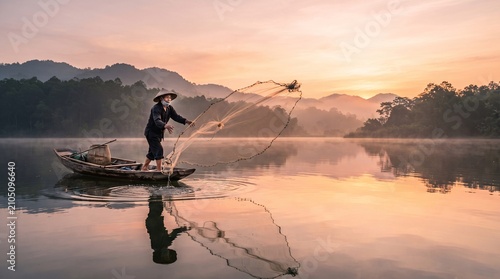 Elderly fisherman wearing traditional hat casts a large circular net from a wooden boat during a beautiful golden misty sunrise