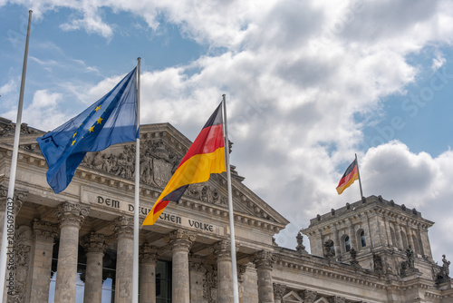 European Union and German flags wave before the Reichstag facade. Classical columns, sculpture and clouds form a strong image of European and national government.