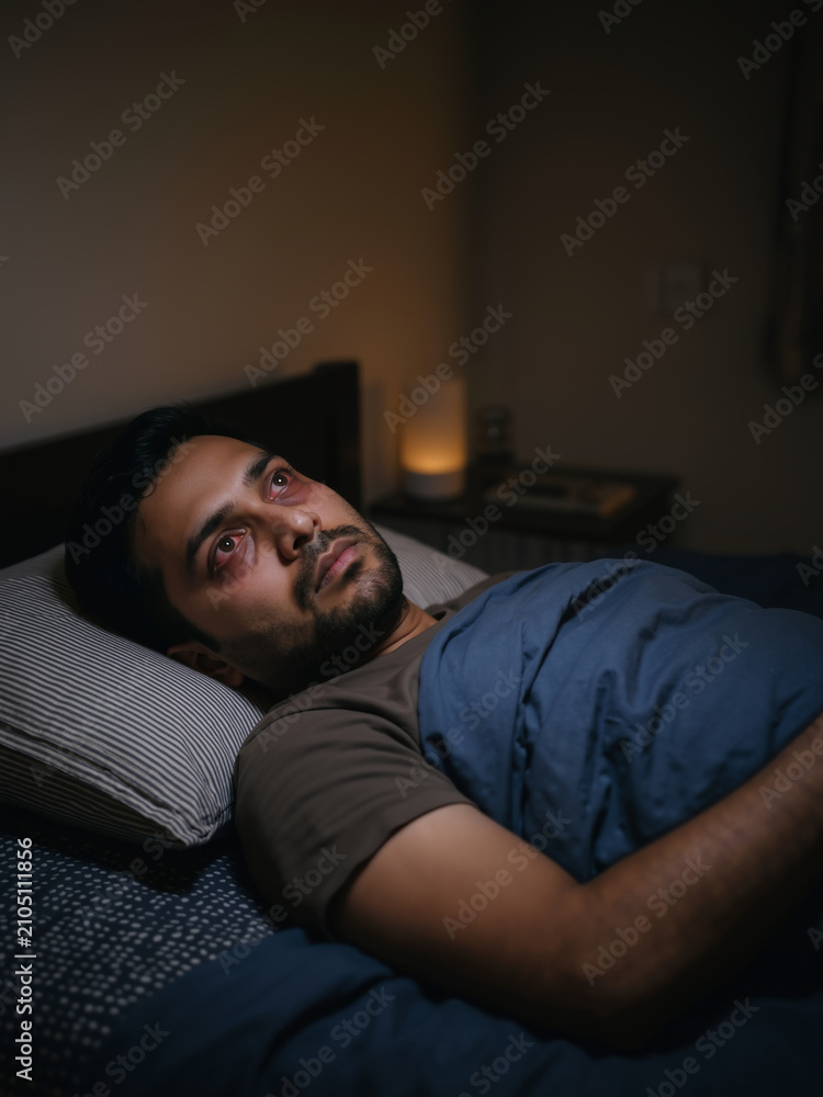 Obraz premium South asian man lying awake in bed at night with red bloodshot eyes. Sleeplessness and insomnia health issues vertical banner with copy space