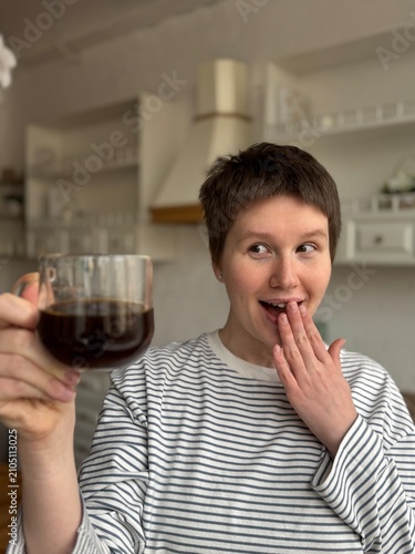 Woman holding coffee cup and covering mouth with, in kitchen, with coffee cup, striped shirt, kitchen cabinets, from a close-up perspective, with white, gray accents.