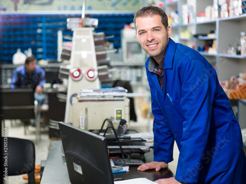 Foto Portrait of a smiling male worker in a blue uniform leaning over a desk with a computer in a hardware store warehouse, surrounded by stocked shelves and retail inventory