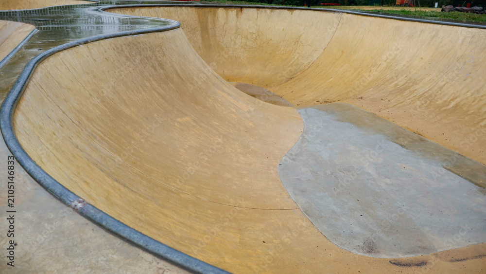 Obraz premium Concrete skatepark bowl showcasing its empty, curved interior with wet surfaces and metal coping, reflecting the aftermath of a rainy day in an outdoor urban sports zone