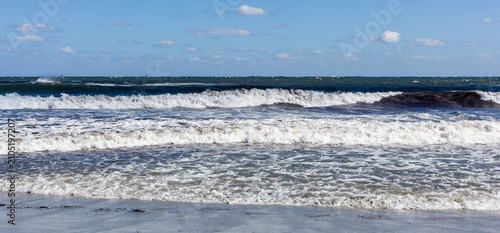 Waves crash on sandy beach in Sardinia, Italy. Calm sea under blue sky with clouds. Coastal shoreline with gentle waves. Mediterranean seascape during summer.