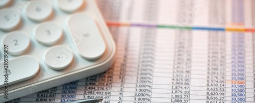 White calculator rests beside colorful financial data sheets. Numbers ripple across rows in soft focus, hinting at analysis or planning. No people appear, so emotions are not applicable