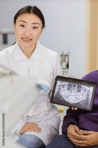 A female dentist in a white coat shows a patient a digital dental X-ray on a tablet screen, discussing treatment options