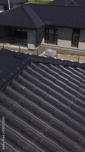 Vertical video. Aerial view moving along the black metal tile roof of a modern house under construction, detailing the material, gutter system, and snow guards on a sunny day with distinct shadows