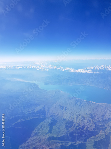 Spectacular aerial view of rugged mountains, a turquoise glacial lake, and snow-capped peaks photographed from an airplane at cruising altitude. The clear blue sky and dramatic landscape create a peac